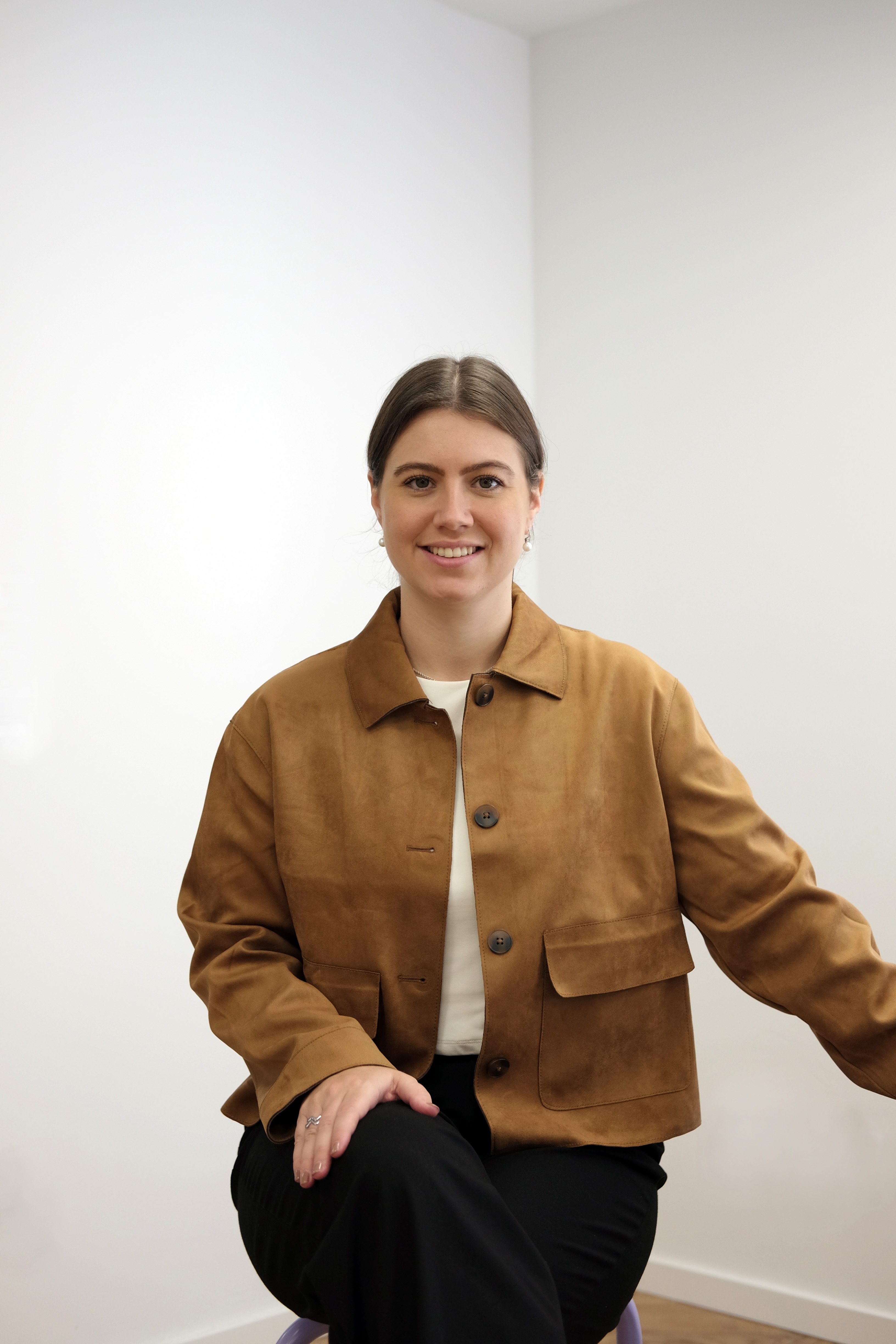 Lisa-Marie in brauner Jacke sitzt auf einem Stuhl in einem hellen Raum und schaut in die Kamera